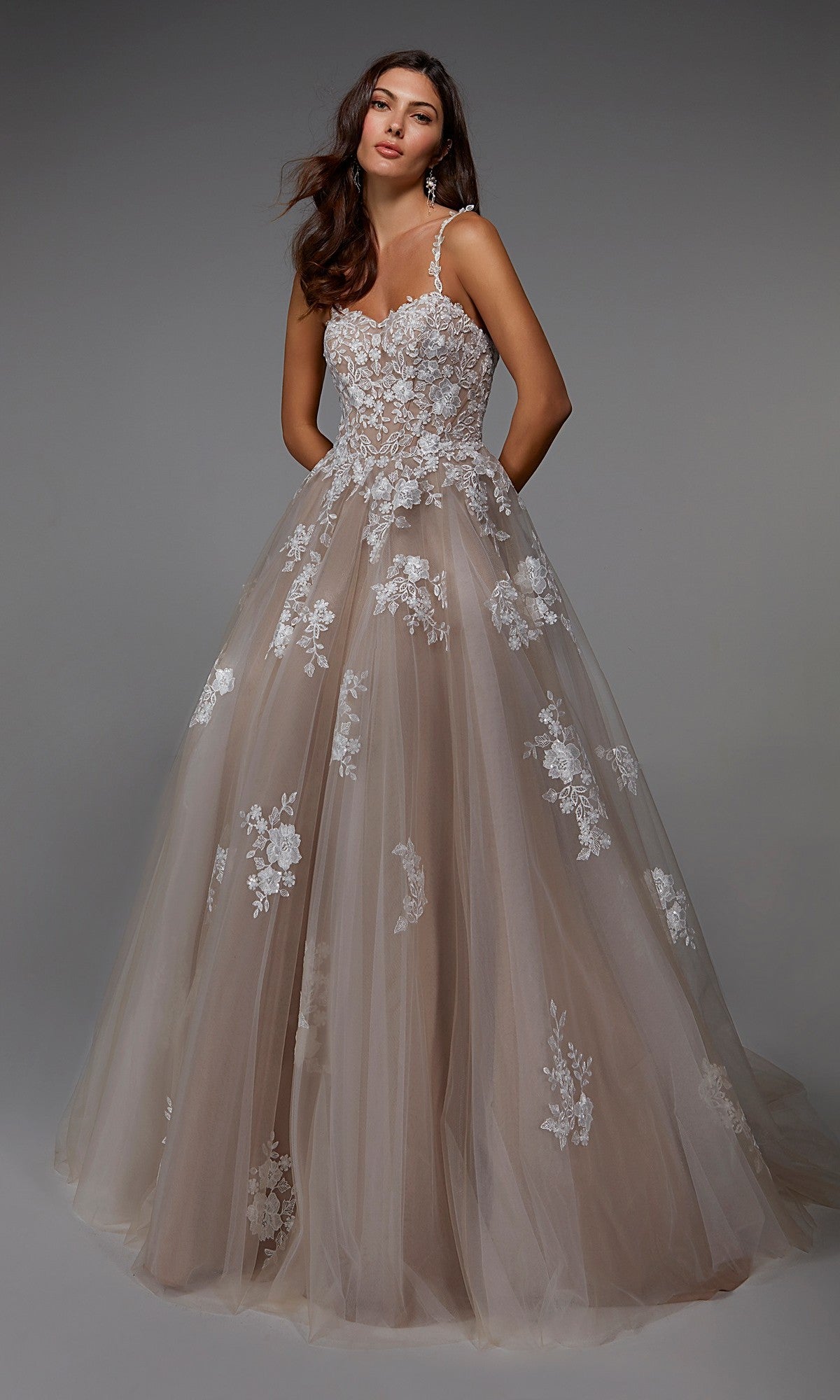 A woman poses gracefully against a plain gray background wearing the Alyce 7043, a long formal blush tulle ballgown with white floral lace appliqué and thin straps.